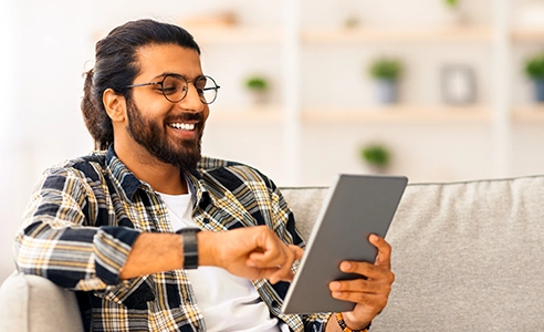 Relaxed young indian man chilling at home with digital tablet, sitting on couch, copy space.